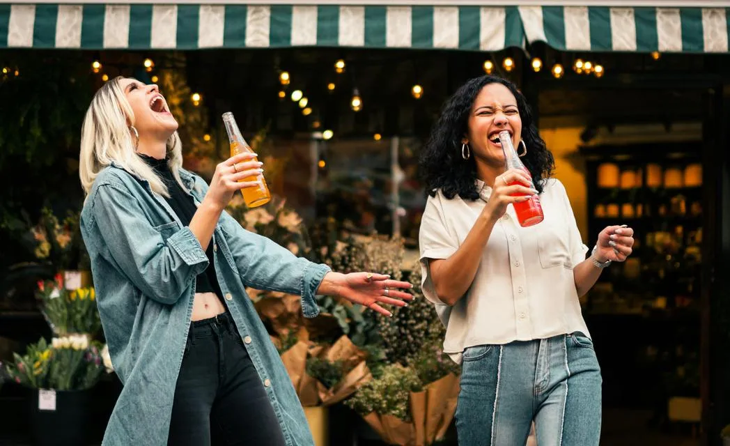 Deux amies riant, bouteilles à la main, devant une terrasse
